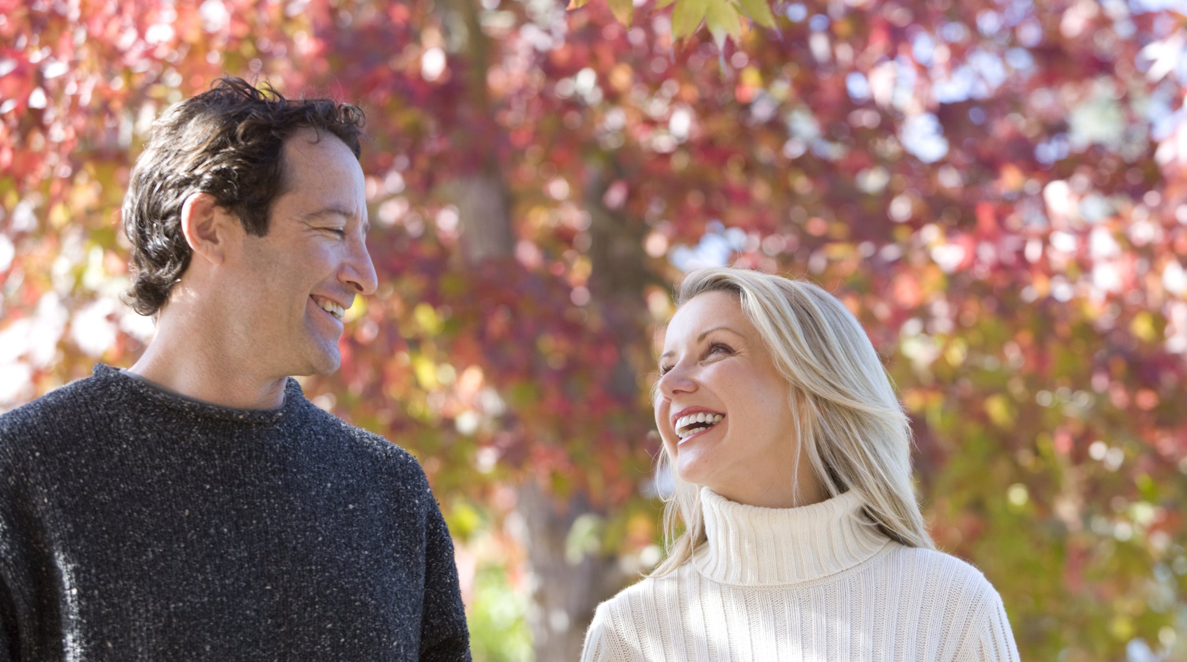 Man and woman smiling at each other in front of autumn trees with colorful leaves